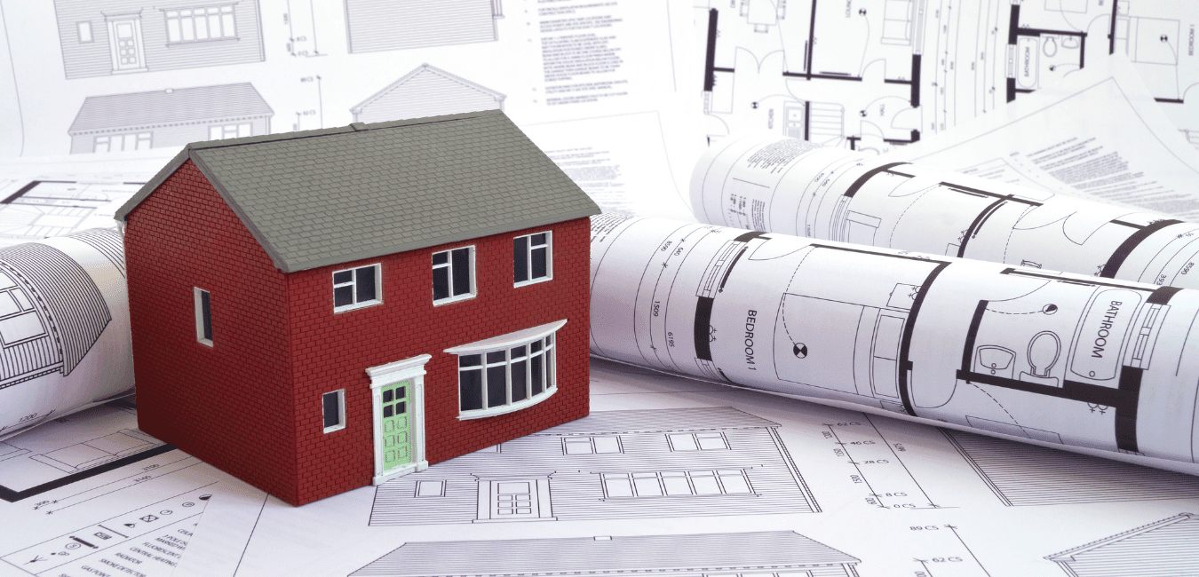 A model of a red brick house with a green door sits atop rolled and flat architectural blueprints and floor plans, symbolising the first steps in Managing Your Self Build.
